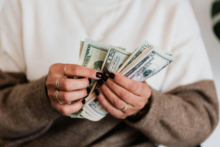 Pessoa segurando cédulas de dólar nas mãos enquanto conta os valores. Suas unhas estão pintadas de preto e seus dedos adornados com anéis dourados.