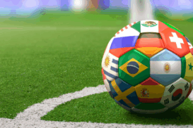Bola de futebol posicionada em um campo, próxima à linha do escanteio. O destaque está no design da bola, composta por bandeiras de diversos países. Ao fundo, vê-se parte do estádio desfocado, com arquibancadas coloridas e iluminação suave.