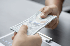 Pessoa entregando notas de dólar a outra em um ambiente de escritório. Sobre a mesa, há documentos com gráficos, um laptop e uma calculadora