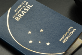 Passaporte brasileiro azul-marinho apoiado sobre uma mesa escura, ao lado de óculos, fones de ouvido e um caderno verde.
