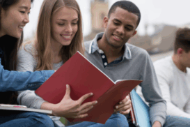 Grupo de jovens sentados em um gramado, estudando juntos ao ar livre. Eles folheiam livros e sorriem. Ao fundo, há construções históricas que remetem a uma cidade europeia.